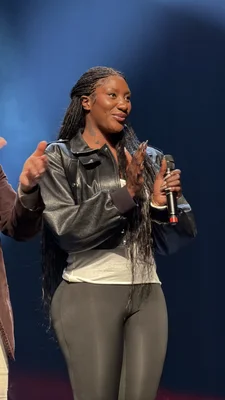 Aya Nakamura en concert, vêtue d'une veste en cuir noire et leggings gris, micro à la main.