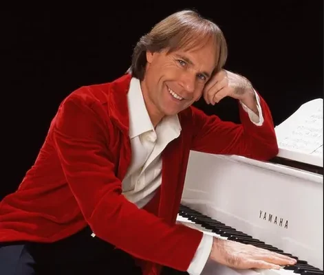 Richard Clayderman en veste rouge, souriant près d'un piano Yamaha blanc.