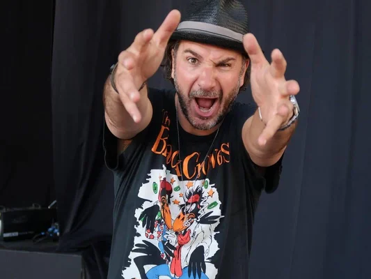 Michaël Youn en chapeau noir et t-shirt graphique, geste expressif vers la caméra.