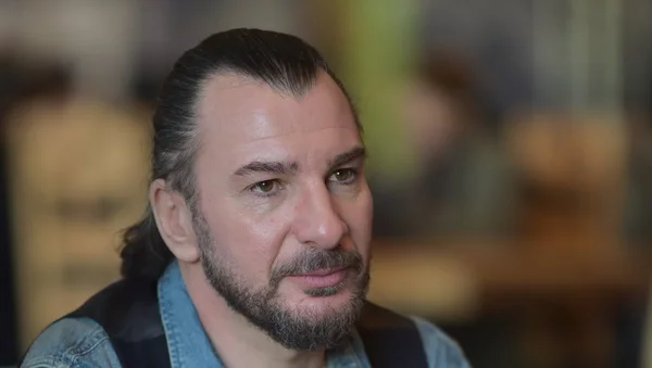Michaël Youn en chemise en jean bleu, cheveux longs attachés.