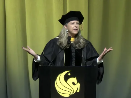 Gloria Caulfield s'adressant aux diplômés de l'UCF depuis le pupitre, les mains levées, lors de la cérémonie de remise des diplômes