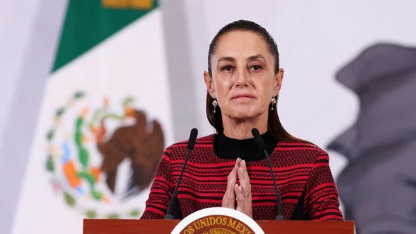 Claudia Sheinbaum, avec des cheveux bruns et portant un haut rouge à motifs, s'exprimant derrière un podium avec deux microphones devant le drapeau mexicain.