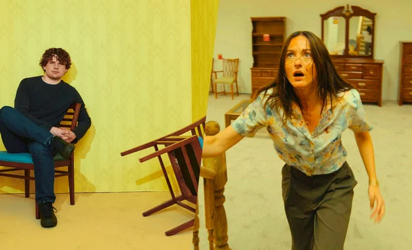 Scène du film Backrooms : tension entre deux personnages dans une pièce jaune au mobilier vintage.