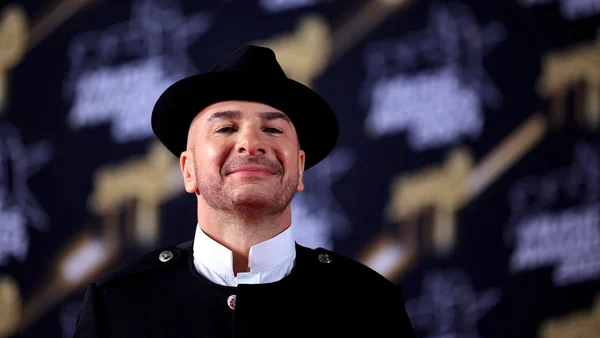 Michaël Youn souriant en chapeau et veste noirs, fond sombre aux reflets dorés.