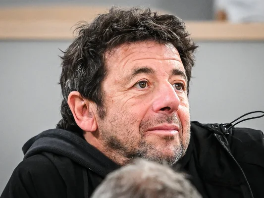 Patrick Bruel, regardant sur le côté, veste à capuche noire.
