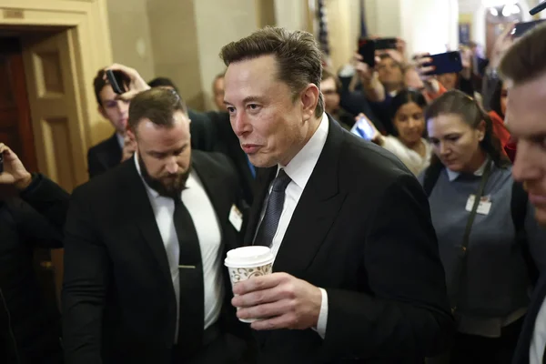 Elon Musk tenant un café, entouré de sécurité et de photographes.