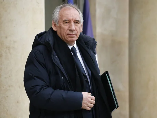 François Bayrou en manteau, tenant un dossier, devant des colonnes.