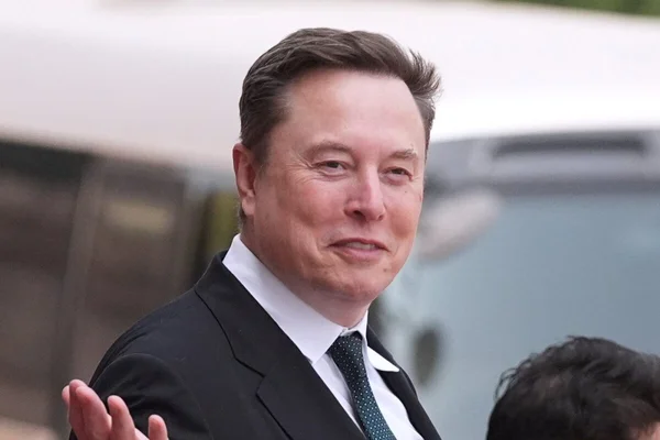 Elon Musk en costume, gesticulant, sur fond flou.