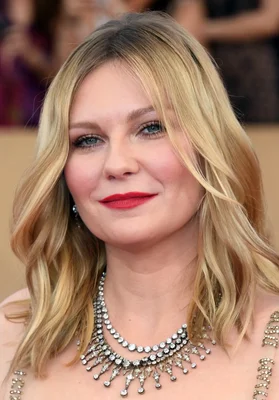 Portrait rapproché de l'actrice Kirsten Dunst, cheveux blonds ondulés, rouge à lèvres rouge, collier de diamants, fond flou.