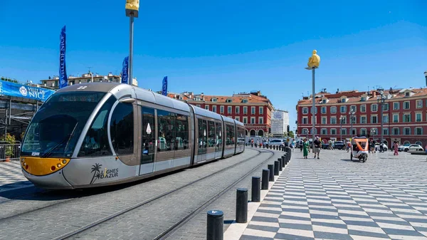 Un tramway moderne traversant une place ensoleillée à Nice, avec des bâtiments rouges et des sculptures jaunes en arrière-plan.