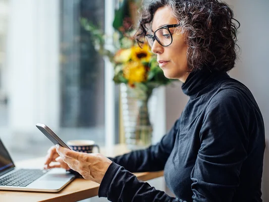 Femme utilisant son smartphone et un ordinateur portable dans un cadre de travail.