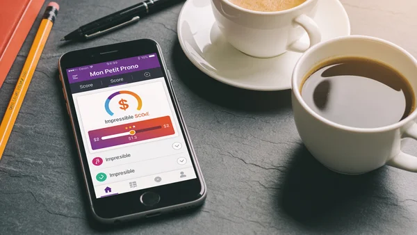 Gros plan sur un smartphone posé sur un bureau, écran affichant l'interface de Mon Petit Prono avec un score improbable, tasses de café et stylos autour, lumière naturelle de bureau