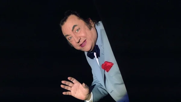 Raymond Devos en costume bleu clair, nœud papillon et pochette rouge, penché vers l'avant la main tendue.