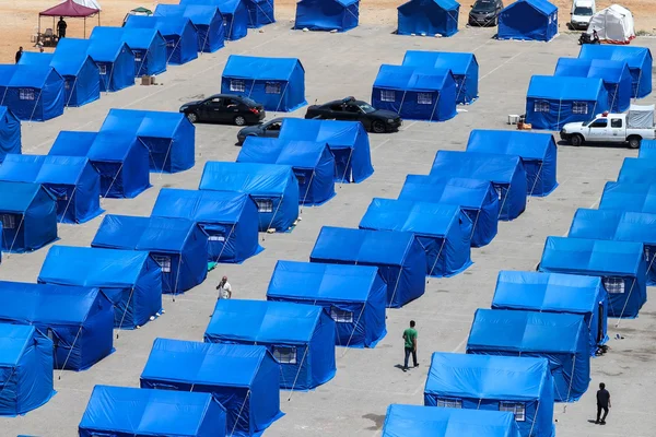 Un campement de tentes bleues installé sur une surface pavée, avec des véhicules et des personnes visibles.