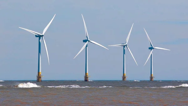 Quatre éoliennes en mer sous un ciel bleu, avec des vagues à la base.