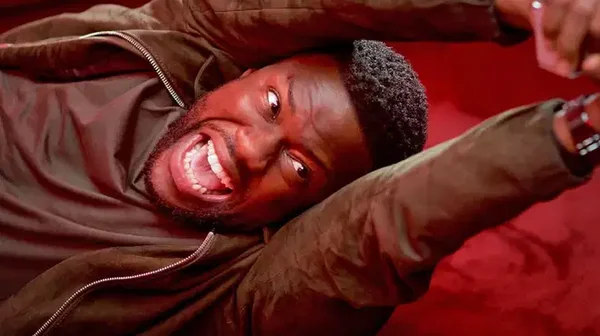 Kevin Hart en plan serré, allongé sur fond rouge, mimique expressive.
