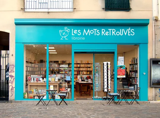 Façade de la librairie 'Les Mots Retrouvés' avec sa devanture turquoise et ses tables en terrasse.