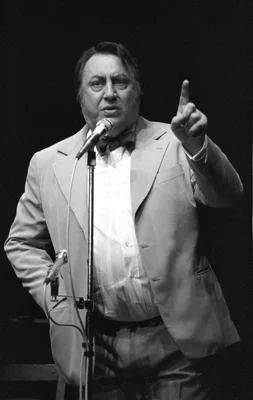 Raymond Devos sur scène, vêtu d'un costume clair et d'un nœud papillon, l'index levé devant un micro.