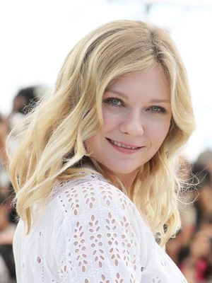 Photographie de l'actrice Kirsten Dunst aux cheveux blonds ondulés, souriant doucement dans un chemisier blanc à œillets, fond flou.