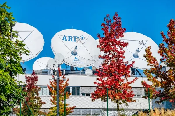 Antennes paraboliques sur un toit, dont une portant le logo ARD, avec des arbres aux couleurs automnales.
