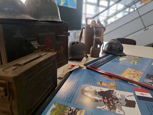 Objets d'époque exposés : casques, munitions et masque à gaz, accompagnés de brochures sur le Débarquement.
