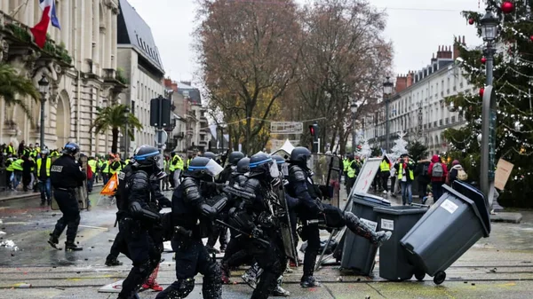 Affrontement entre policiers anti-émeute en tenue complète et manifestants en gilets jaunes dans une rue ; un officier renverse une grande poubelle. Décoration de Noël visible en arrière-plan.
