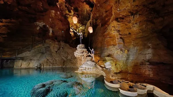 Grotte souterraine avec une formation rocheuse et des statues illuminées, entourée d'eau turquoise.