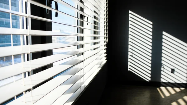 Fenêtre de studio avec volet roulant baissé, soleil extérieur frappant le store, pièce plongée dans la pénombre, angle intérieur