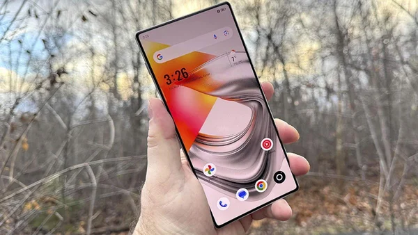 Main tenant un smartphone Samsung affichant son écran d'accueil, avec un arrière-plan flou d'arbres.