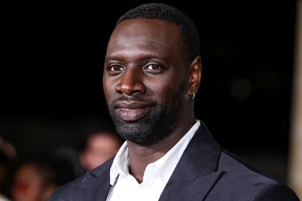Omar Sy en costume sombre, regard de côté, fond sombre.