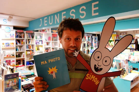 Un libraire présentant un album jeunesse et un personnage de Simon le lapin dans une librairie.