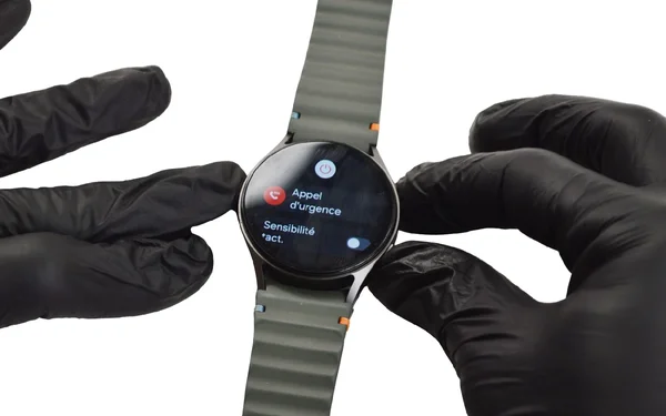 Mains gantées tenant une montre connectée, écran affichant les options 'Appel d'urgence' et 'Sensibilité tact.'.