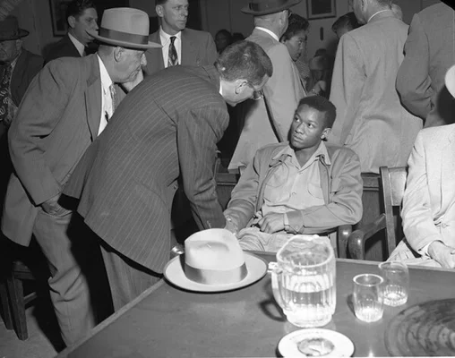 Scène d'audience ou de réunion dans une salle comble, avec un homme en costume rayé s'adressant à un jeune homme noir assis.