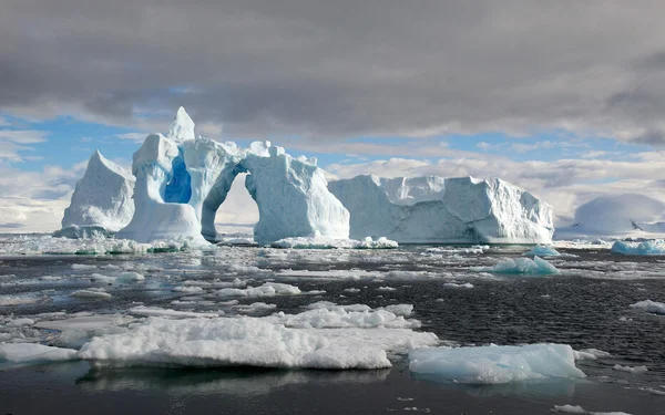 Iceberg sculpté avec une arche naturelle, entouré de glace de mer sous un ciel nuageux.