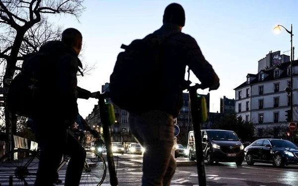 Deux personnes roulant en trottinette électrique à un carrefour de Bruxelles au crépuscule.
