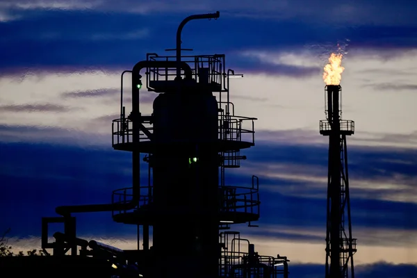Installation industrielle, probablement une raffinerie, avec une torche de gaz brûlant au crépuscule.