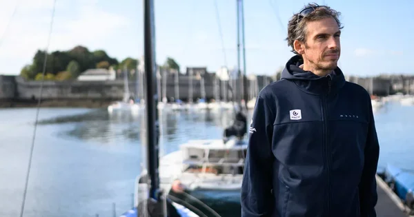 Charlie Dalin debout dans un port, regardant au loin, avec des voiliers amarrés en arrière-plan.