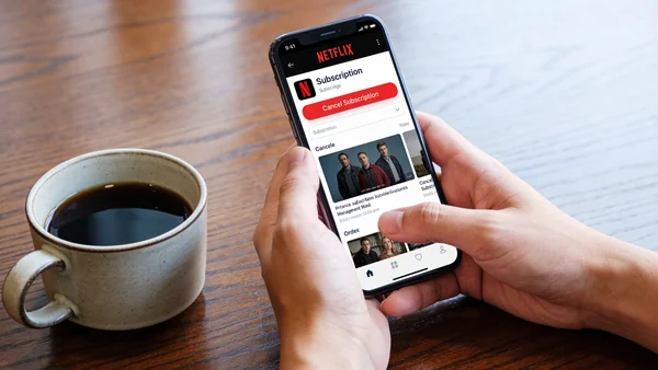 Deux mains tenant un smartphone, l'écran affiche la page de gestion des abonnements Netflix, une tasse de café à côté, ambiance sobre et décidée