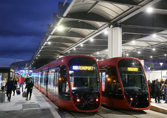 Deux trams rouges de la métropole niçoise à l'arrêt dans une station moderne, au crépuscule, montrant les destinations Aéroport et Magnan.