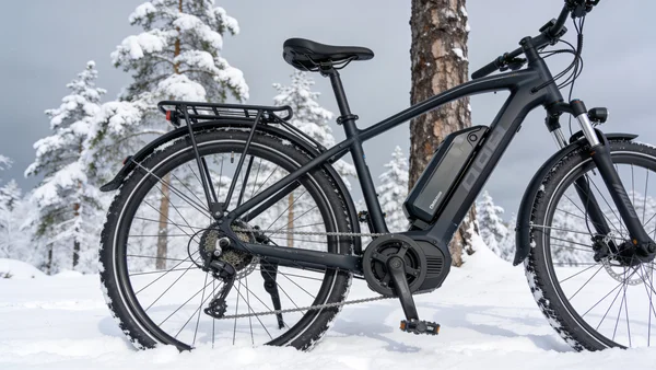 Vélo électrique garé dans la neige, batterie visible sur le cadre, paysage hivernal en arrière-plan, ciel gris et arbres enneigés