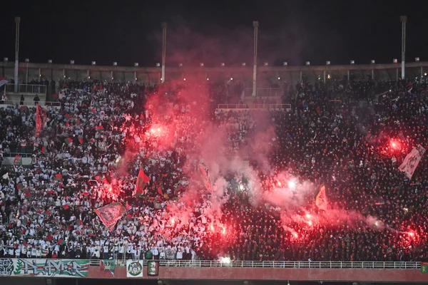 Célébration du titre du MC Alger dans un stade, avec des fumigènes rouges créant une épaisse fumée.
