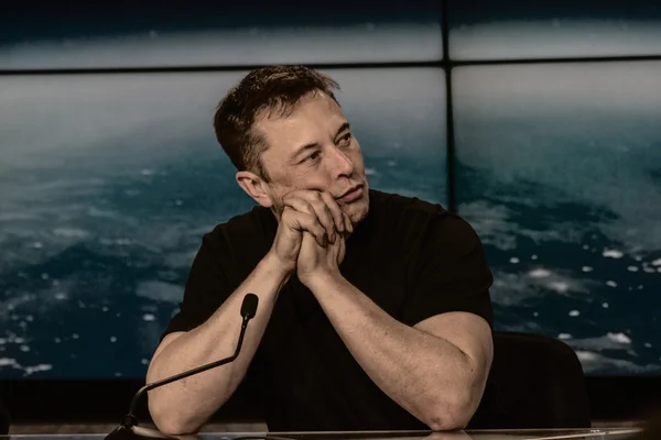 Elon Musk lors d'une conférence de presse, assis à une table avec un micro.