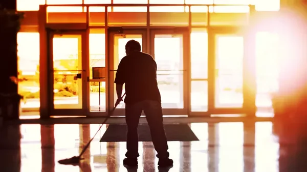 Silhouette d'un agent d'entretien nettoyant un sol devant des portes vitrées, illustrant de manière générique le thème du travail et des aides sociales.