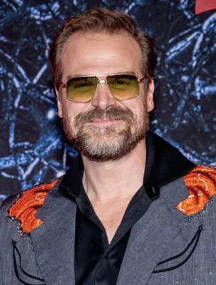 David Harbour dans une pose élégante avec lunettes aviateur et veste brodée.