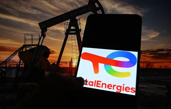 Smartphone affichant le logo de TotalEnergies devant une pompe à pétrole au crépuscule.