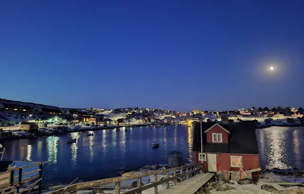 Ville côtière groenlandaise illuminée sous la lune, avec des maisons colorées et un port.