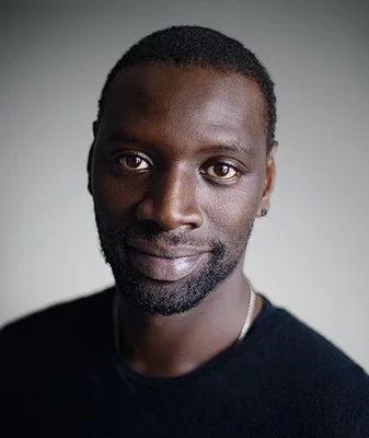 Omar Sy en portrait studio, chemise noire sur fond gris.