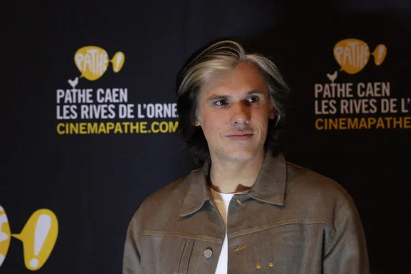 Orelsan devant l'écran Pathé Caen, veste marron et chemise blanche.