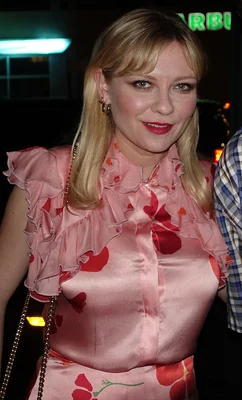 Photographie en plan moyen de Kirsten Dunst, frange blonde, chemisier floral rose à volants, rouge à lèvres, fond sombre avec enseigne au néon.
