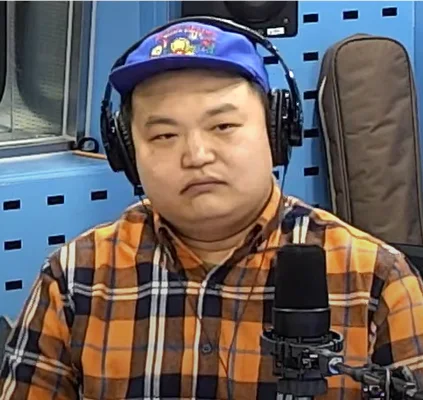 Ko Kyu-pil en studio de radio, casquette et casque audio, devant un micro.